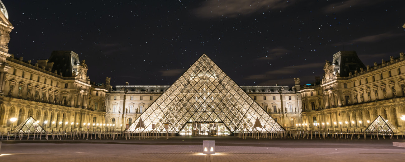 Trinagular glass building of the famous Louvre Museum centred, with buildings illuminated with lighting and stars prominent in the night sky above